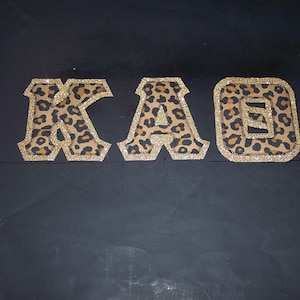 May include: Three decorative letters spelling "KAO" are displayed against a dark background. Each letter features a leopard print pattern with a gold glitter border. The letters are likely intended for crafting or personalization.