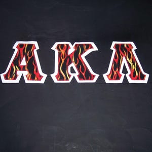 May include: The letters "AKA" are displayed in a bold, stylized font. Each letter is outlined in white and filled with a flame design in red, orange, and yellow against a black background. The overall effect is striking and eye-catching.