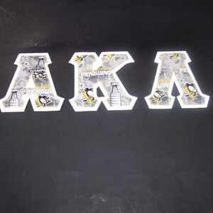 May include: Three white, die-cut letters spelling "AKA" with a Pittsburgh Penguins hockey theme. Each letter is decorated with images of the Stanley Cup, the team logo, and the word "Penguins".