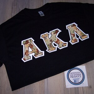 May include: Black t-shirt with the letters "AKA" in a white outline, filled with a camouflage pattern. A sticker with the text "Affinity Licensed Product" is visible.