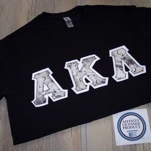 May include: Black t-shirt with the letters "AKA" in a white, textured design. The letters have a celestial pattern with moons and stars. A small sticker with text is in the lower right corner.