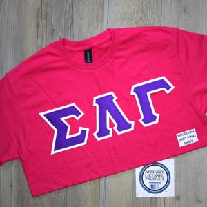 May include: A hot pink short-sleeved t-shirt featuring the Greek letters Sigma, Lambda, and Gamma in purple with a white outline across the chest. A small white rectangular label on the lower right reads HELICONIA (HOT PINK) SHIRT.