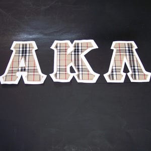 May include: Three fabric letters spelling "AKA" are displayed against a dark background. The letters feature a beige, black, and red plaid pattern with a white border. The letters are cut out and appear to be fabric patches.