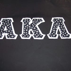 May include: Three white-bordered patches spelling "AKA" are displayed against a black background. The letters are filled with a black and white skull pattern. The patches are likely for clothing or accessories.