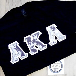 May include: Black t-shirt with the letters "AKA" in a white and gray design. The shirt has a tag that says "Affinity Licensed Product". The shirt is laying on a light wood surface.