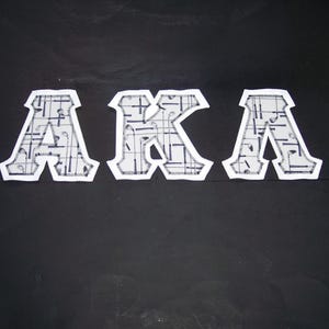 May include: Three white and gray fabric letters spelling "AKA" are displayed against a black background. The letters have a patterned design and are outlined with a white border. The letters are likely patches or appliques.