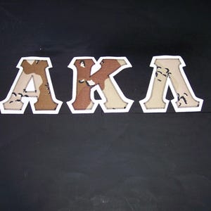 May include: Three fabric letters spelling "AKA" are displayed against a black background. The letters are outlined in white with a stitched edge. The letter shapes are filled with a camouflage pattern in shades of brown and tan.