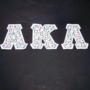 May include: White fabric letters spelling "AKA" with a colorful golf tee pattern. The letters are outlined with a blue stitched border, set against a black background. The design is likely for a sorority or organization.