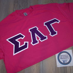 May include: A bright pink t-shirt with the Greek letters ΣΛΓ in a purple glitter design outlined in white. The shirt is lying flat on a wooden surface. A small sticker with a blue and white design is in the lower right corner.