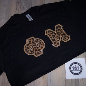May include: Black t-shirt with leopard print letters "ΦM" outlined in gold glitter. A small white sticker with a circular design is in the lower right corner. The shirt is laying on a wooden surface.