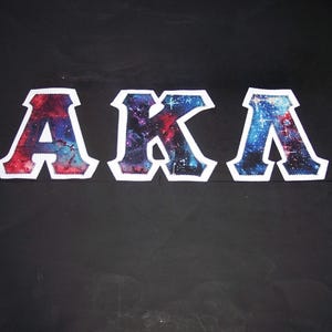 May include: Three embroidered letters, "AKA", are displayed against a black background. The letters are white-bordered and filled with a colorful galaxy design featuring shades of red, blue, and purple. The letters are evenly spaced.