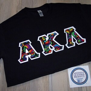 May include: Black t-shirt with the letters "AKA" in a white outline, filled with a colorful skull pattern. The shirt has a crew neck and short sleeves. A white sticker with blue text is in the bottom right corner.