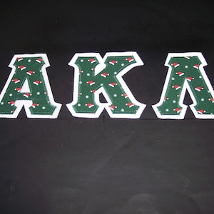 May include: Three large felt letters spelling "AKA" in green with a white outline. The letters are decorated with a pattern of red and white Santa hats.