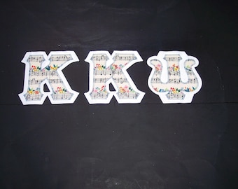 May include: Three fabric letters forming the Greek letters Kappa, Kappa, and Psi. Each letter features a white border and a printed pattern of black musical notes on a cream background, accented with small, colorful floral illustrations.