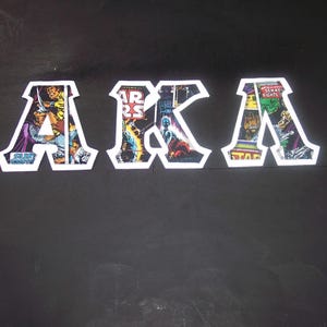 May include: Three white-bordered patches spelling "AKA" with comic book art filling each letter. The letters are arranged horizontally against a dark background. The comic book art features various characters and scenes.