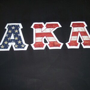 May include: The letters "AKA" are displayed against a black background. The letter "A" is navy blue with gold stars. The "K" and "A" are red and white stripes, resembling the American flag. Each letter has a white border.