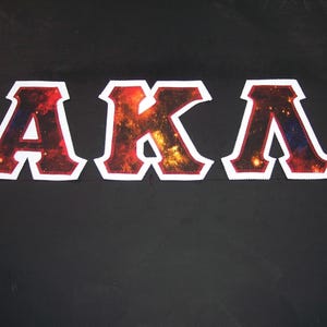 May include: Three embroidered patches spelling out "AKA" in a collegiate font. The letters are filled with a galaxy-like design in shades of red, orange, and yellow, outlined in white with a red border. The patches are displayed on a black background.