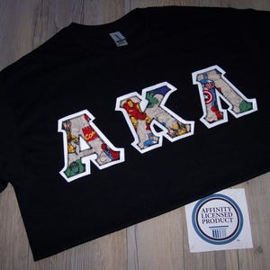 May include: Black t-shirt with the letters "AKA" in a comic book style design. The letters are outlined in white and filled with various comic book characters. A sticker with a blue and white design is in the bottom right corner.