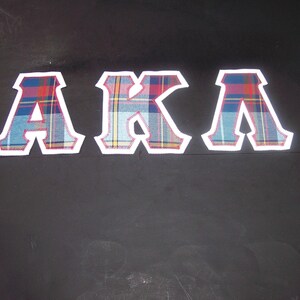May include: Three fabric letters spelling "AKA" are displayed against a dark background. Each letter is outlined in white and filled with a plaid pattern in shades of blue, red, and yellow. The letters are evenly spaced.