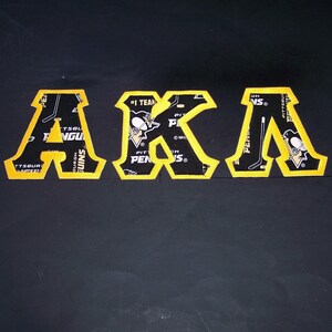 May include: Black and yellow embroidered letters spelling "AKA". The letters are in a block style with a yellow border. The letters are filled with a black and white pattern with the words "Pittsburgh Penguins" and hockey-related imagery.