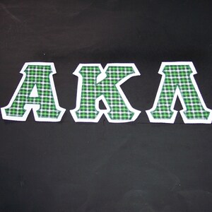 May include: Three embroidered letters, "AKA", are displayed against a black background. The letters are white with a green and white plaid pattern. The letters have a white border.