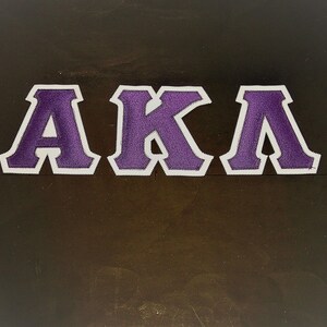 May include: Embroidered patch featuring the letters AKA in purple with a white border. The letters are in a bold, blocky font. The patch is likely for apparel or accessories.