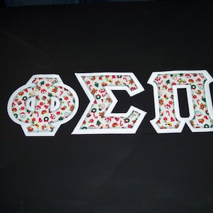 May include: Three Greek letters, Phi, Sigma, and Pi, are displayed on a black background. Each letter is white with a patterned fabric interior featuring Christmas-themed designs. The letters are stitched with a white border.