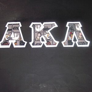 May include: White-bordered letters spelling "AKA" with Star Wars themed fabric filling. The fabric features characters and symbols from the Star Wars franchise. The letters are set against a dark background.