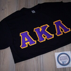 May include: Black t-shirt with the letters "AKA" in purple with a gold outline. The shirt is on a wooden surface, and a sticker that says "Affinity Licensed Product" is in the bottom right corner.