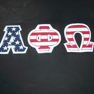 May include: Three fabric Greek letters, Alpha, Phi, and Omega, featuring a patriotic design. The Alpha has a navy blue background with white stars, while the Phi and Omega display red and white horizontal stripes. Each letter has a white border.