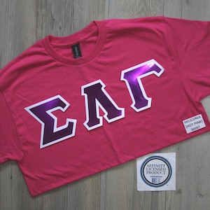 May include: A hot pink short-sleeved t-shirt featuring the Greek letters Sigma, Lambda, and Gamma in a metallic purple finish with a white border. A small white label reads HELICONIA (HOT PINK) SHIRT and an AFFINITY LICENSED PRODUCT circular logo.