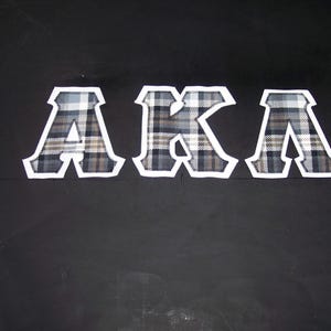 May include: The letters "AKA" are displayed in a plaid pattern with a white border against a black background. The letters are in a collegiate style, with a bold, blocky font.