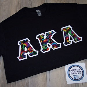 May include: Black t-shirt with a white outline of the letters "AKA" in a colorful, skull-patterned design. The shirt is a short-sleeved crew neck style.