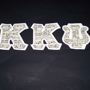 May include: Three fabric letters spelling KKU, each featuring a vintage beige sheet music print with black musical notes and staves. The letters have a distinct white border and a zigzag stitched edge, standing out against a dark background.