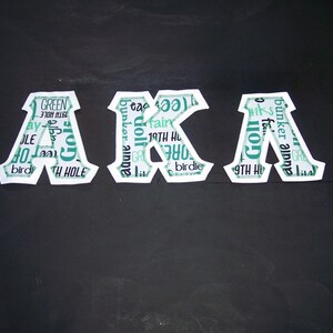 May include: Three white fabric letters spelling "AKA" with a golf-themed pattern in teal and white. The letters are on a black surface. The pattern includes golf-related words like "golf", "birdie", and "19th hole".