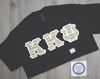 May include: A black short-sleeved t-shirt featuring the Greek letters KKU in a large, block-style font. The letters are filled with a vintage-style musical notation pattern and outlined in white. A small label reads BLACK SHIRT and AFFINITY LICENSED PRODUCT.
