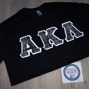 May include: Black t-shirt with the letters "AKA" in a white outline with a plaid pattern. The shirt has a crew neck and a small sticker in the bottom right corner that says "Affinity Licensed Product".