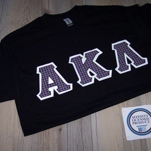 May include: Black t-shirt with the letters "AKA" in white outlined, purple and white plaid design. A small white sticker with blue text and a barcode is in the lower right corner. The shirt is laying on a wooden surface.