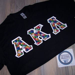 May include: Black t-shirt with the letters "AKA" in white, each filled with a colorful dice pattern. The shirt is on a wooden surface, with a sticker in the bottom right corner.