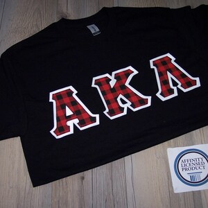May include: Black t-shirt with the letters "AKA" in a red and black plaid pattern outlined in white. The shirt has a crew neck and short sleeves. A white sticker with a blue border and text is in the bottom right corner.