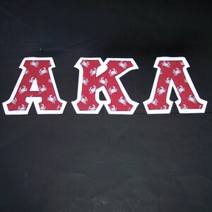 May include: Three decorative fabric letters spelling "AKA". The letters are red with a white crab pattern and white borders. The letters are displayed against a black background.
