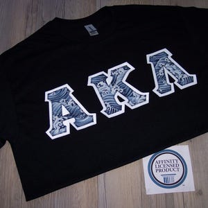 May include: Black t-shirt with the letters "AKA" in a white outline, filled with a blue and white wave pattern. A sticker with the text "Affinity Licensed Product" is visible.