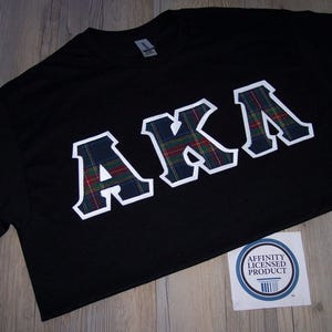 May include: Black t-shirt with the letters "AKA" in white outlined, plaid-filled block letters. A sticker with the text "Affinity Licensed Product" is in the lower right corner. The shirt is displayed on a wooden surface.