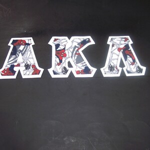 May include: White-bordered patches spelling "AKA" with a Spider-Man theme. The letters are filled with a pattern featuring Spider-Man, webs, and a red, white, and blue color scheme. The patches are set against a black background.