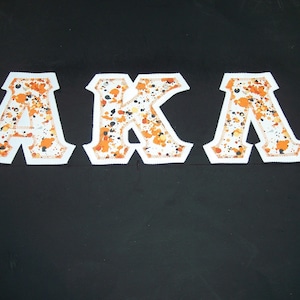 May include: Three white fabric letters spelling "AKA" with orange and black splatter designs. The letters are outlined with a white border and stitched edges, set against a black background.