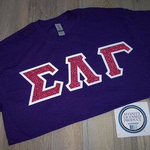 May include: A purple t-shirt with the Greek letters ΣΛΓ in a red glitter design outlined in white. The shirt has a tag and a sticker that says "Affinity Licensed Product".