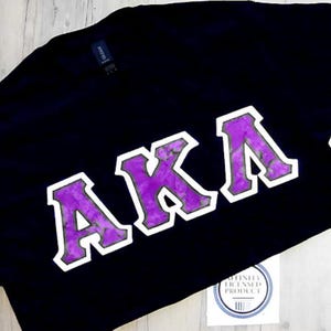 May include: Black t-shirt with the letters "AKA" in a purple and white design. The letters have a marble-like pattern and are outlined in white. The shirt is on a light wood surface.