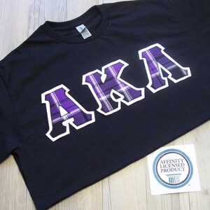 May include: Black t-shirt with the letters "AKA" in white outlined purple plaid. The shirt is made of a soft material and is likely a casual wear item. A sticker with the text "Affinity Licensed Product" is visible.