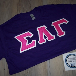 May include: Purple t-shirt with a pink Greek letter design that says "ΣΛΓ".