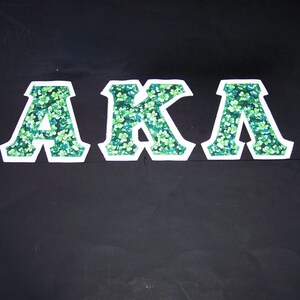 May include: Three white-bordered letters, "AKA", are displayed against a black background. The letters are filled with a pattern of green and light green clover-like shapes. The overall design is bold and eye-catching.